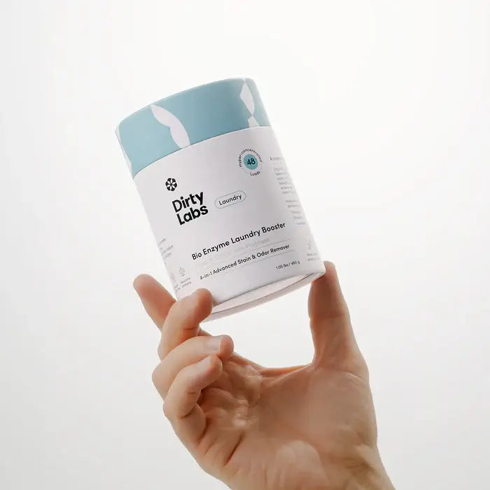 Hand holding a container of 'Dirty Labs' laundry booster against a white background