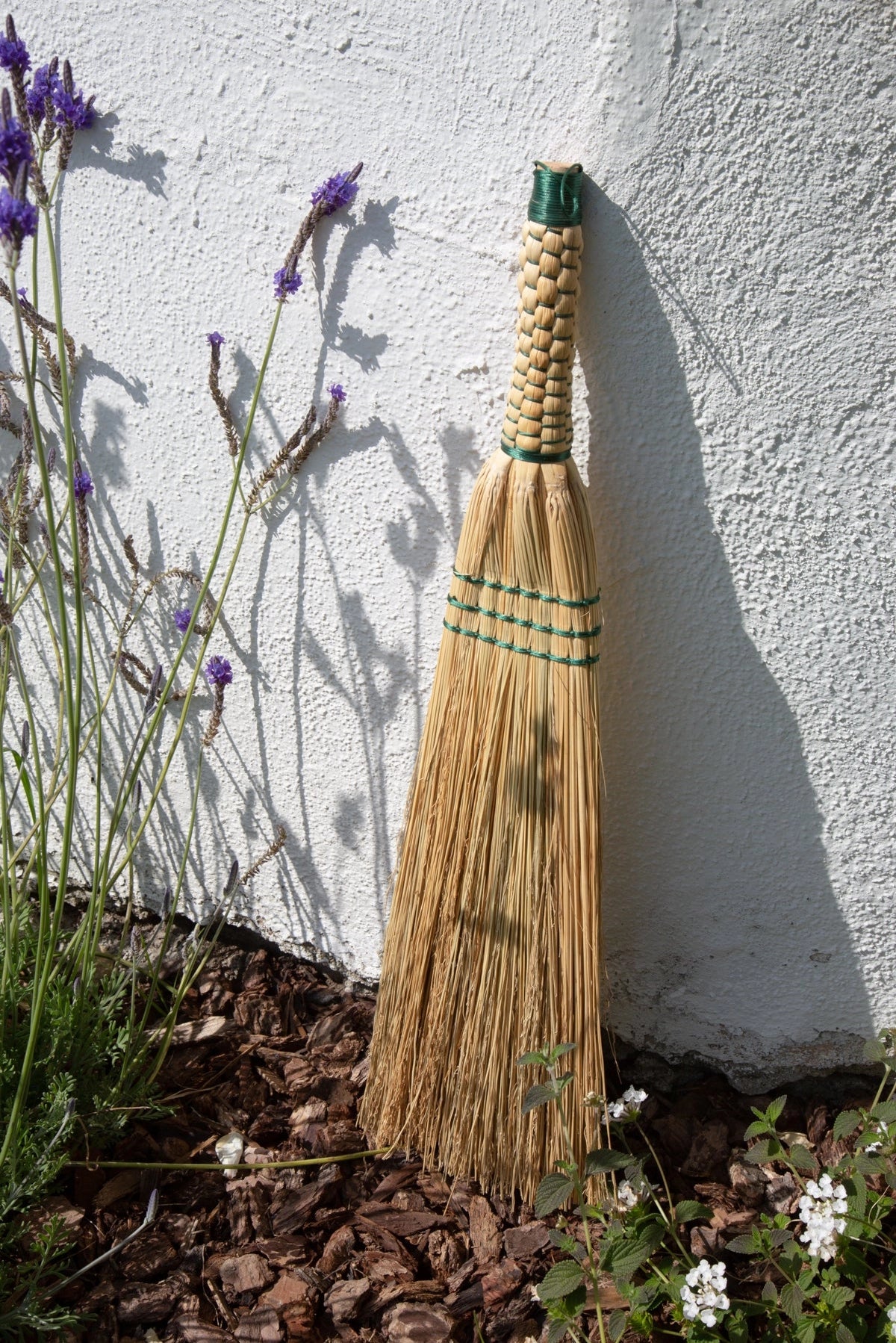 Handmade broom leaning against a white wall with lavender plants.