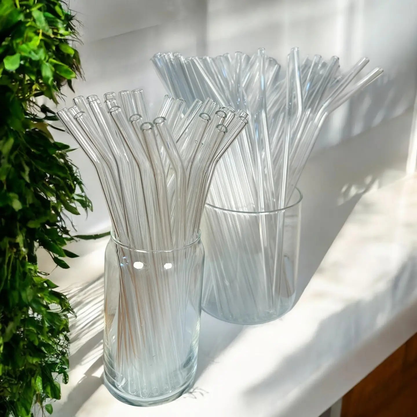 Clear glass containers filled with clear straws on a white surface with a plant in the background.