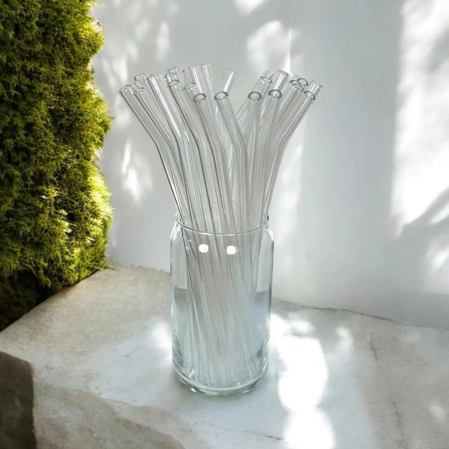 Clear glass vase filled with clear glass straws on a light surface with a blurred green plant in the background.