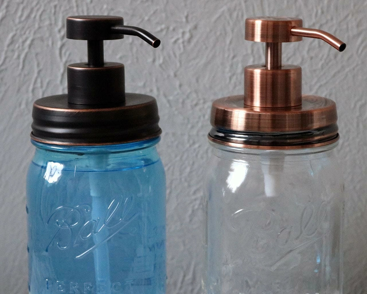 Two glass jars with metallic pump tops, one blue and one clear, on a textured gray background.